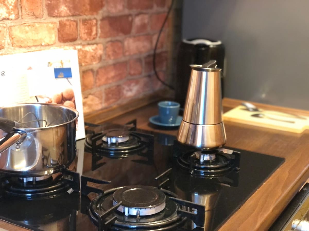 A well-equipped kitchen corner features a stainless steel pot on a gas stovetop, accompanied by a modern coffee maker and a small blue cup. Dark wooden countertops and exposed brick walls provide a stylish backdrop to this functional cooking area.
