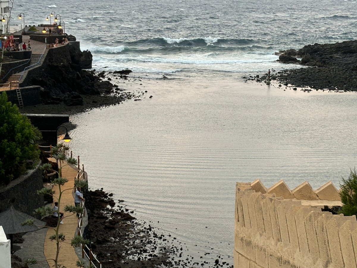 A serene coastal view depicts gentle waves lapping at the rocky shoreline. A path lined with green foliage leads towards the water, while visitors can be seen along the promenade. The atmosphere reflects tranquility, with the ocean and shore merging harmoniously.