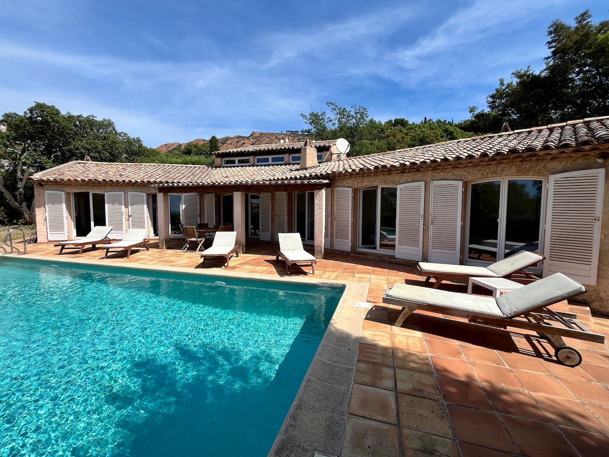 The outdoor area features a private pool surrounded by several sun loungers. Large glass doors and windows allow natural light to fill the space, while the rustic terracotta tiles provide a warm touch. Lush greenery and scenic views can be seen in the background.