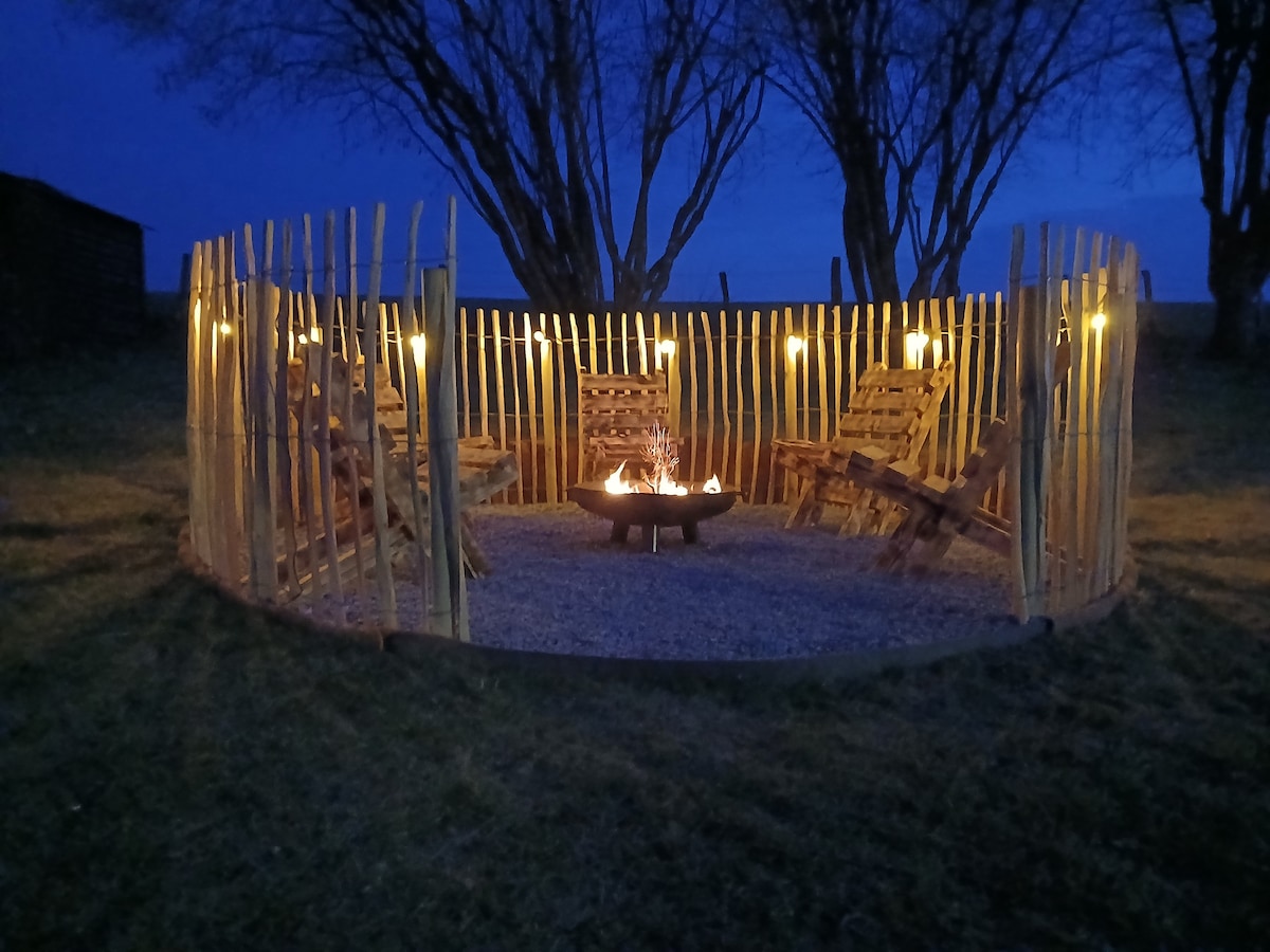 An outdoor seating area is enclosed by a circular arrangement of wooden posts, providing a rustic ambiance. Several wooden chairs surround a central fire pit. Soft lighting is fixed to the posts, casting a warm glow in the evening setting.