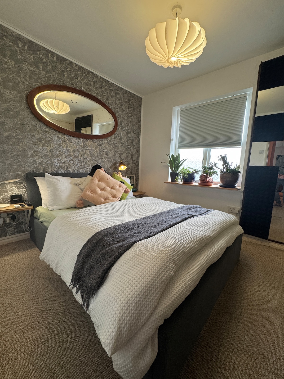 A bright sleeping area is presented with a spacious bed featuring soft bedding and a decorative throw. A round mirror reflects light from a cozy pendant lamp. Potted plants adorn the windowsill, complementing the textured wallpaper and creating a serene atmosphere.