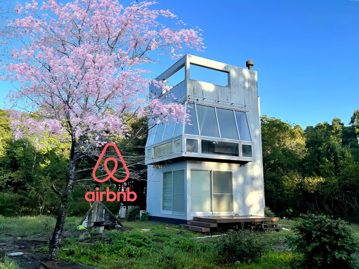 A modern three-story house is surrounded by greenery, featuring large windows that allow natural light to fill the interior. A blossoming cherry tree is positioned nearby, adding a touch of nature, while a wooden deck at the base provides a space for outdoor relaxation.
