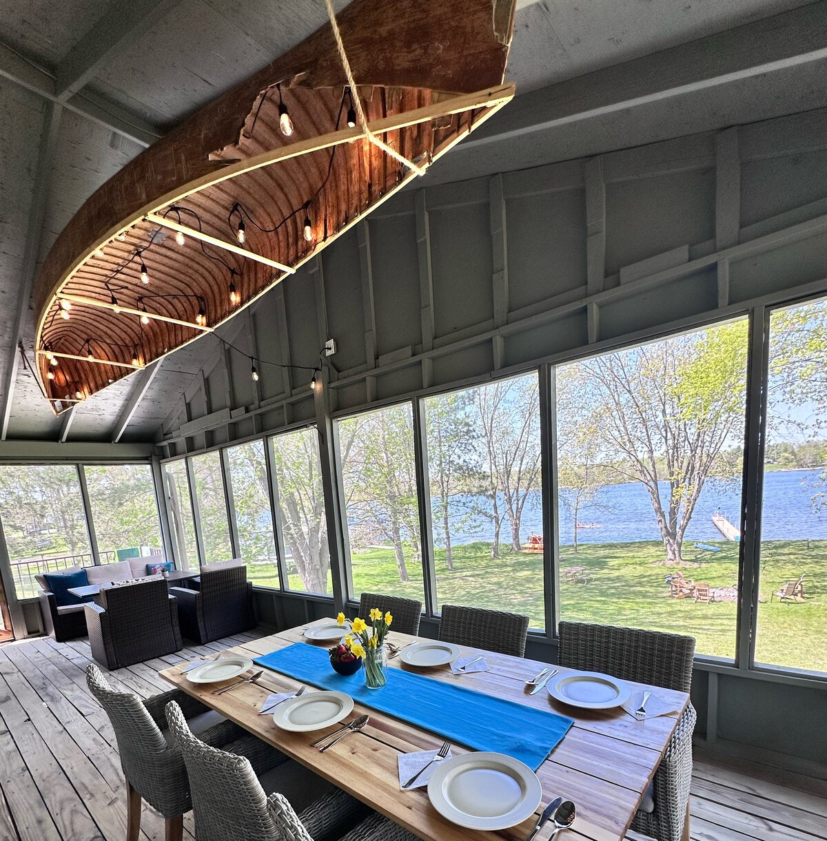 A dining area is surrounded by large windows offering views of the lake. A wooden table is set with plates and a vase of flowers at the center. A cozy seating arrangement with wicker chairs is visible in the background, complemented by a decorative canoe fixture above.
