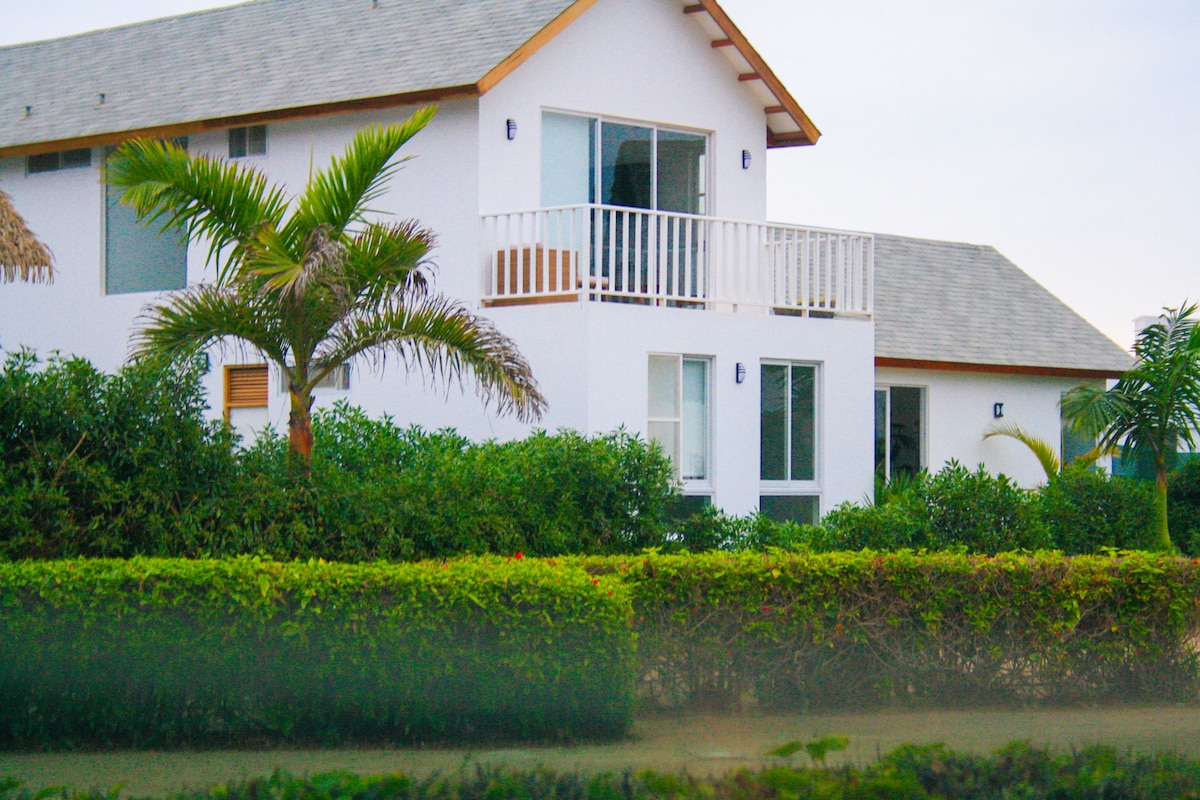 A two-story house is viewed from a distance, featuring a front balcony and large windows that allow natural light to illuminate the interior. Lush greenery surrounds the home, enhancing the tranquil setting. The roof is gently sloped, and a palm tree is visible nearby.