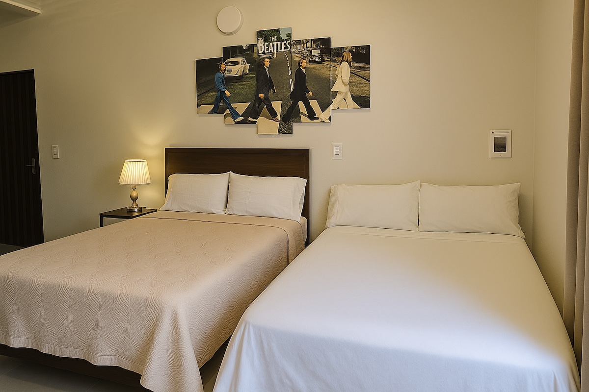 A spacious sleeping area features two beds covered with neutral-toned bedding. A bedside table with a lamp sits beside the larger bed. Artwork depicting a famous band is displayed above the beds, adding a touch of character to the room.
