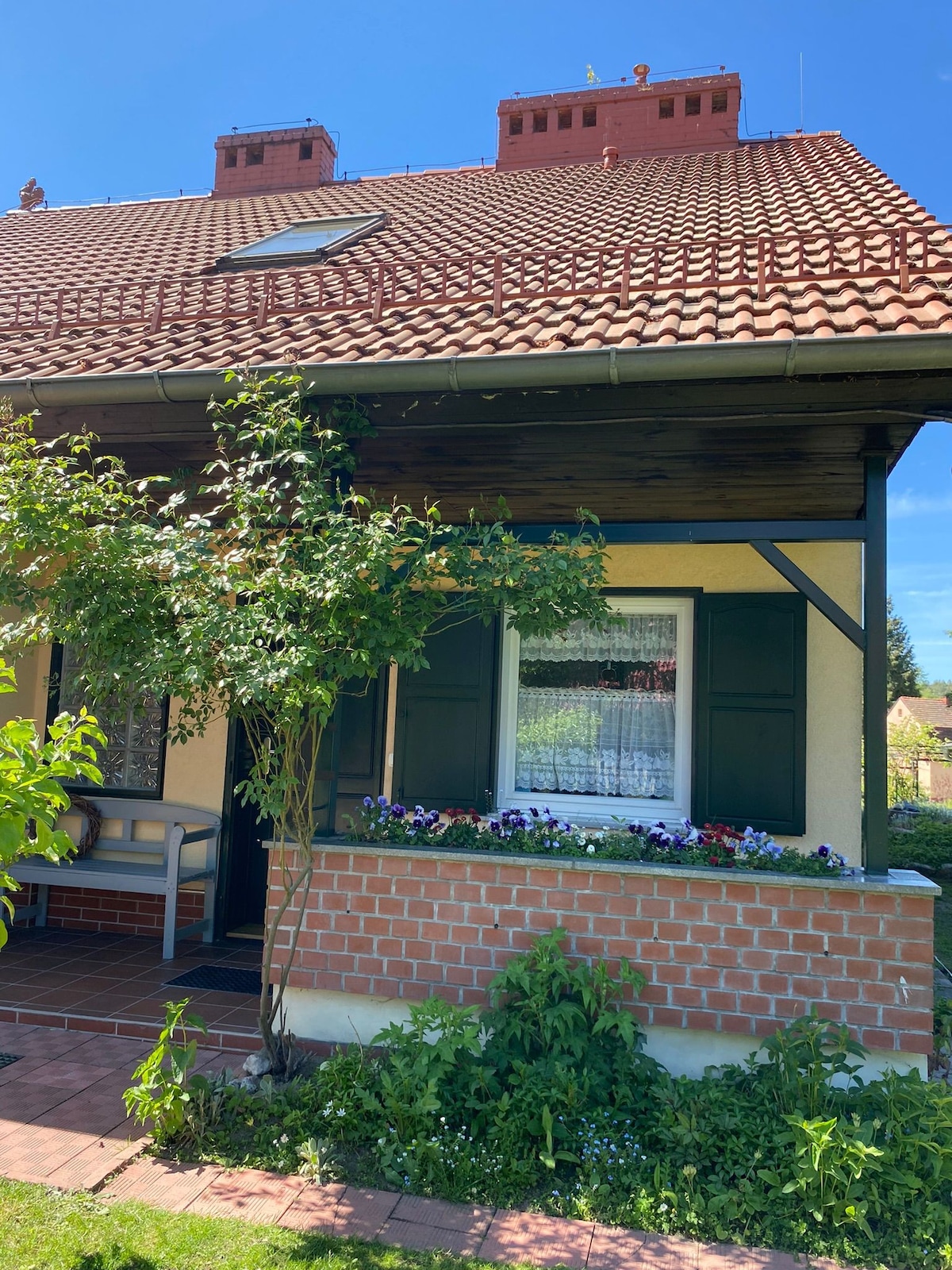 The exterior of a charming house is visible, featuring a sloped red-tiled roof with prominent dormer windows. Green shutters adorn the windows, and a small flower bed displays a variety of colorful blooms beneath the main window. Lush greenery frames the entrance.