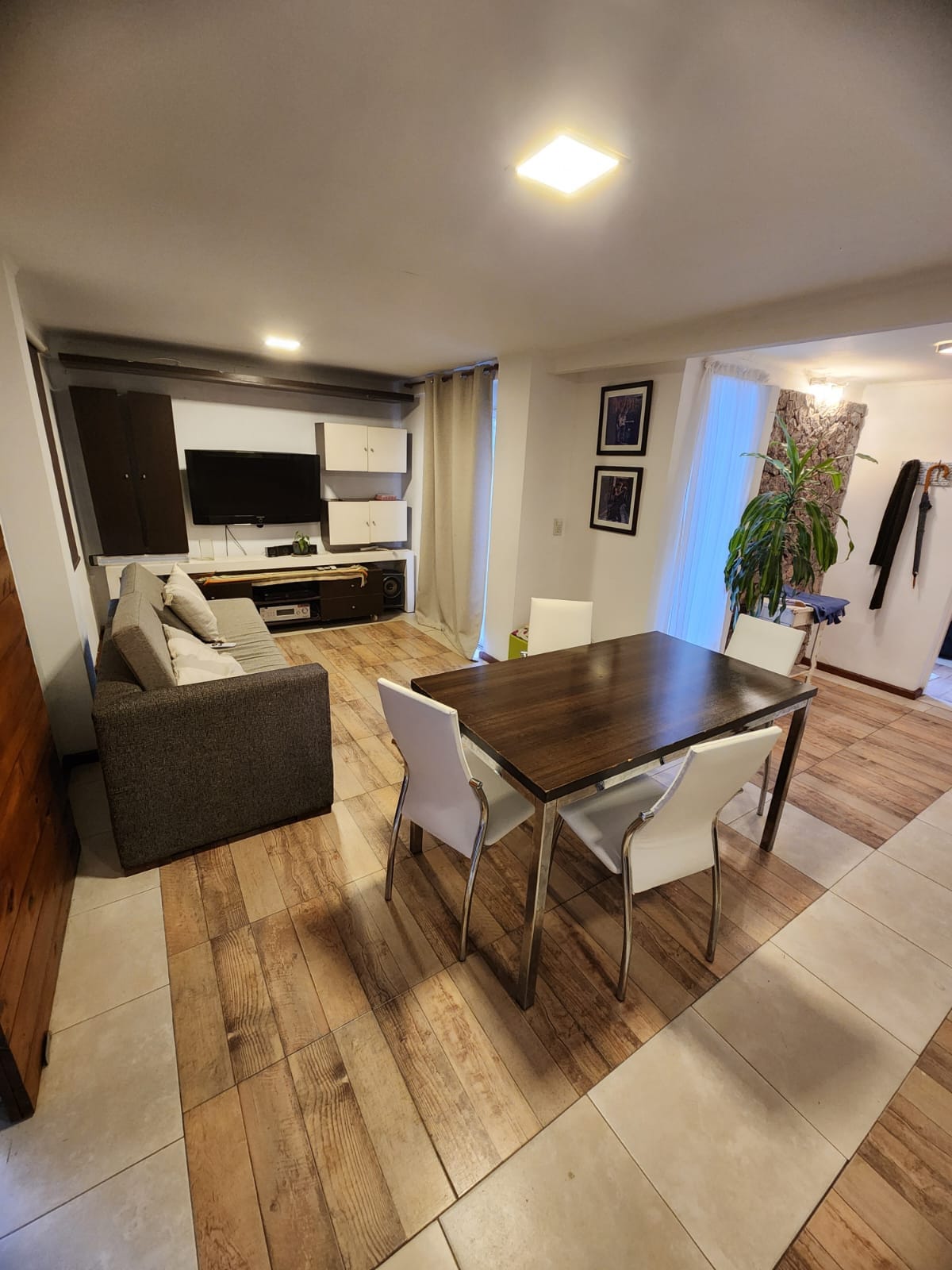 A well-lit living area features a comfortable gray sofa and a dining table with four white chairs. A flat-screen TV is mounted on the wall, with natural light filtering in through a nearby window, creating a bright and airy atmosphere.