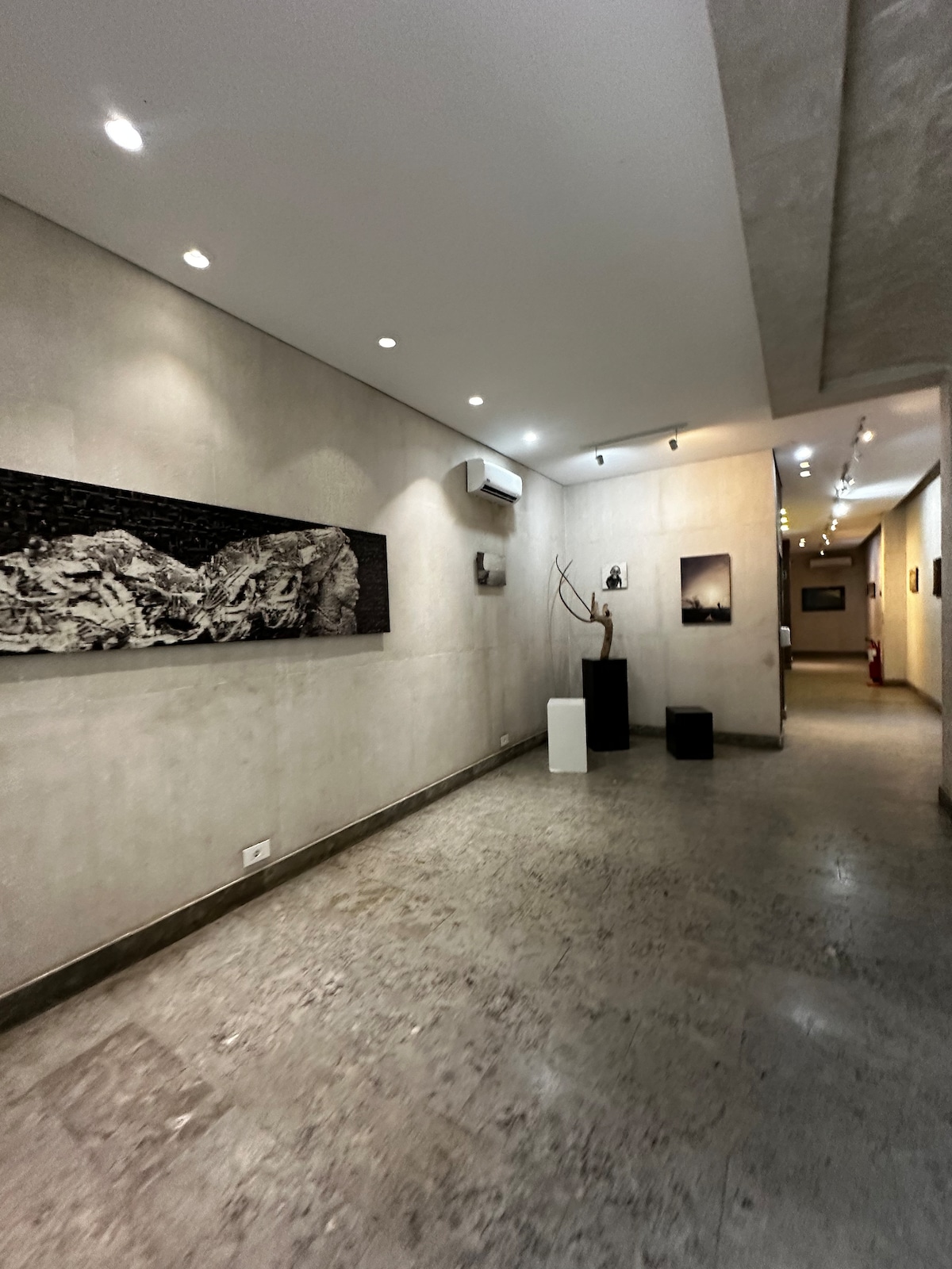 A well-lit hallway features polished stone flooring and textured walls. Black and white artwork is displayed on the left side, while light fixtures provide soft illumination throughout. The space leads to additional rooms, creating a sense of openness and flow.