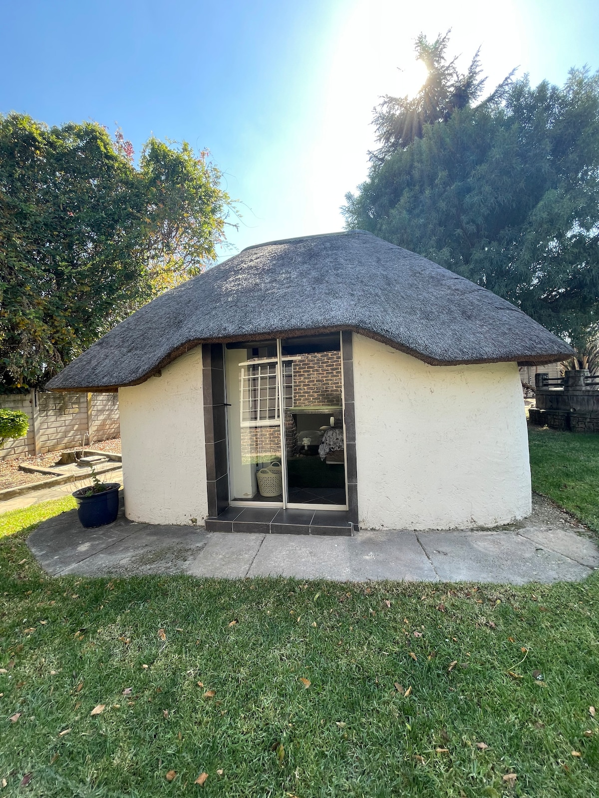 A round structure with a traditional thatched roof is highlighted in this image. The white walls contrast with the natural greenery surrounding the building. A stone pathway leads to the entrance, where large glass doors provide access to the interior.