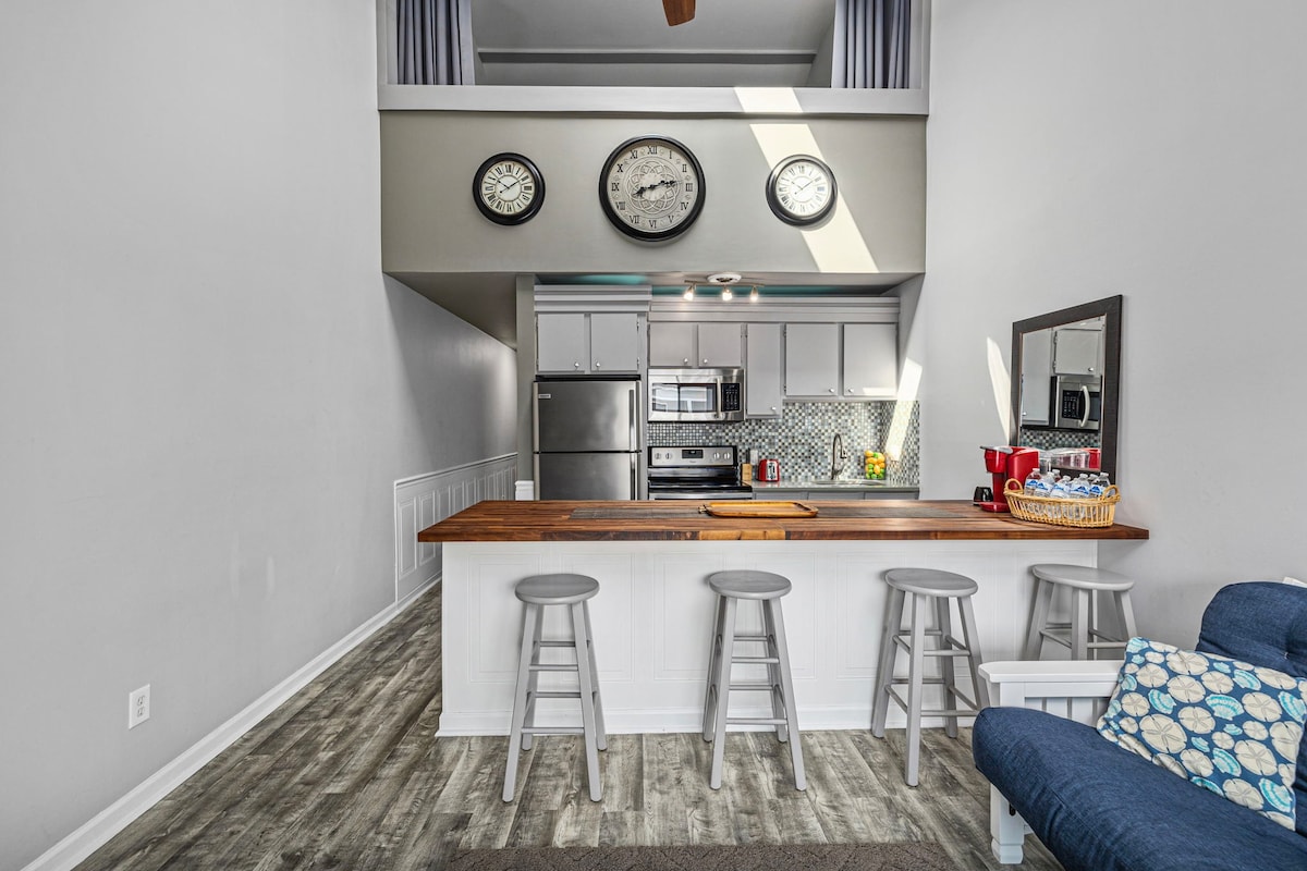 A bright kitchen area features a spacious wooden countertop with four stools positioned along one side. Modern appliances, including a refrigerator and microwave, are visible. Three clocks hang above, indicating different times, while natural light enters through the upper windows, enhancing the open space.