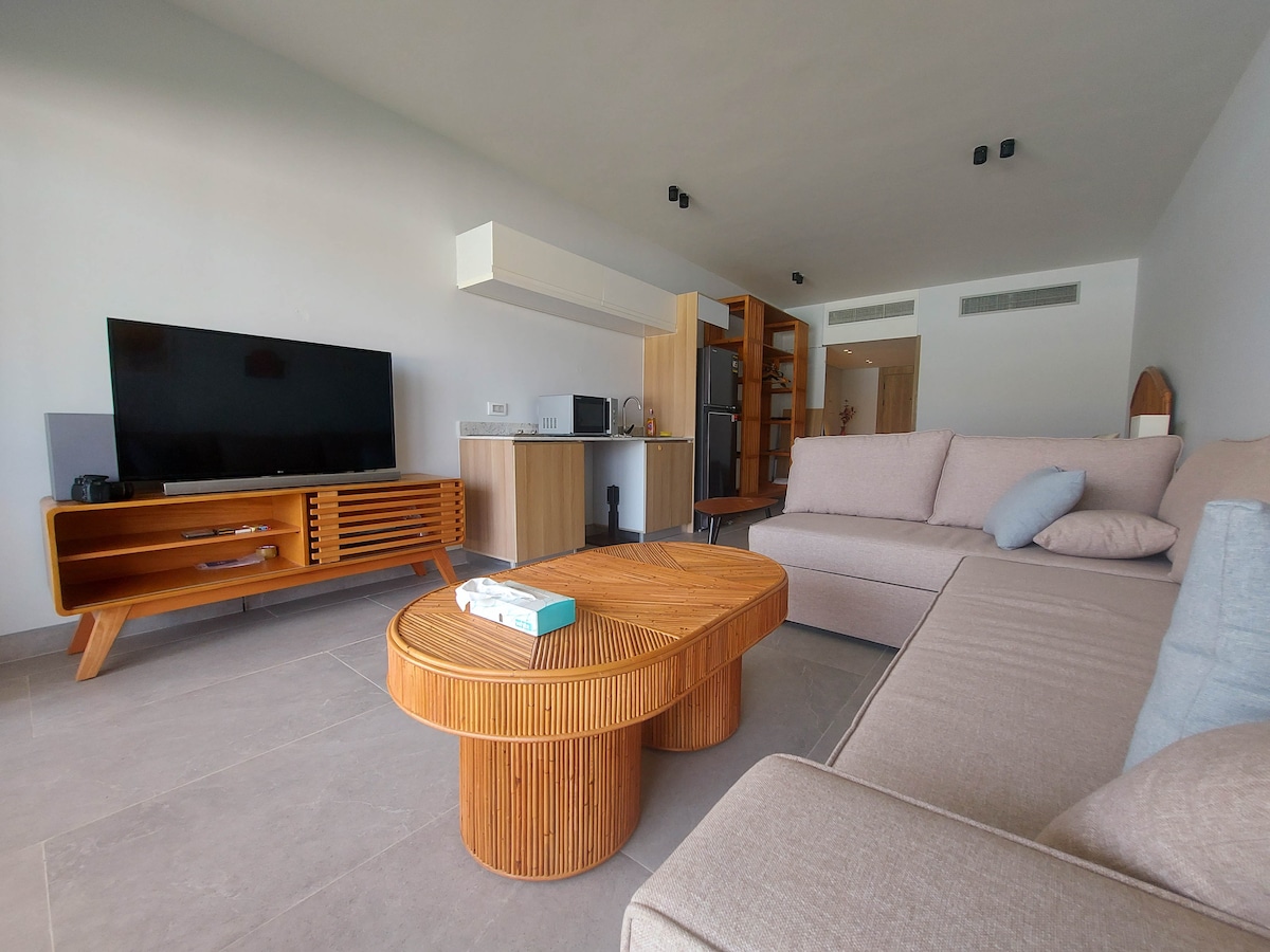 The living area features a light-colored sofa arranged beside a round wooden coffee table. A flat-screen television is mounted on the opposite wall, with a compact kitchenette visible in the background, showcasing modern appliances amid a neutral color palette.