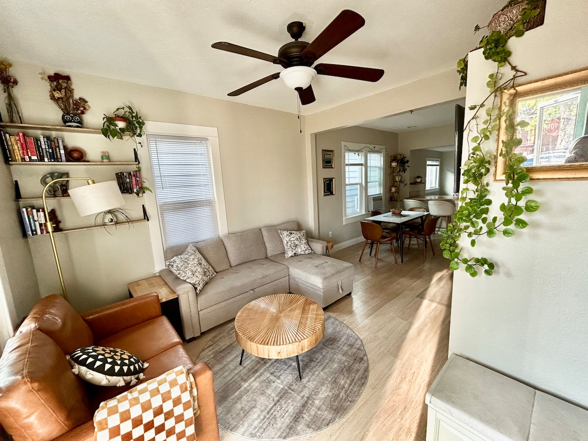 A cozy living area is presented with a light-colored sofa and a brown leather chair. A round coffee table sits in the center, surrounded by a circular rug. Natural light is filtered through nearby windows, enhancing the inviting ambiance.