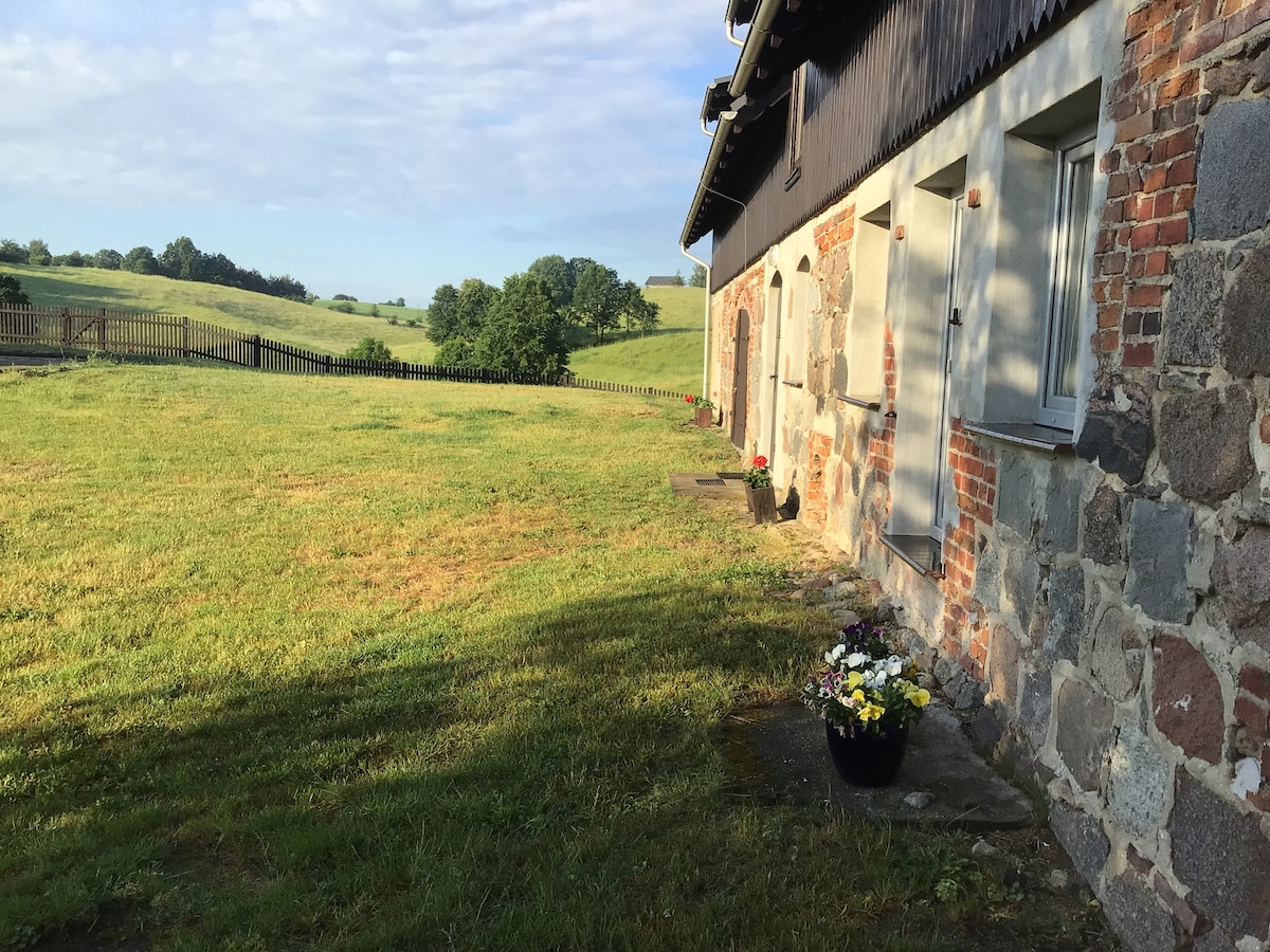 The image shows a grassy area beside a stone and wooden building, with flower pots displayed near the entrance. Rolling hills are visible in the background, along with trees and a wooden fence that outlines the property.