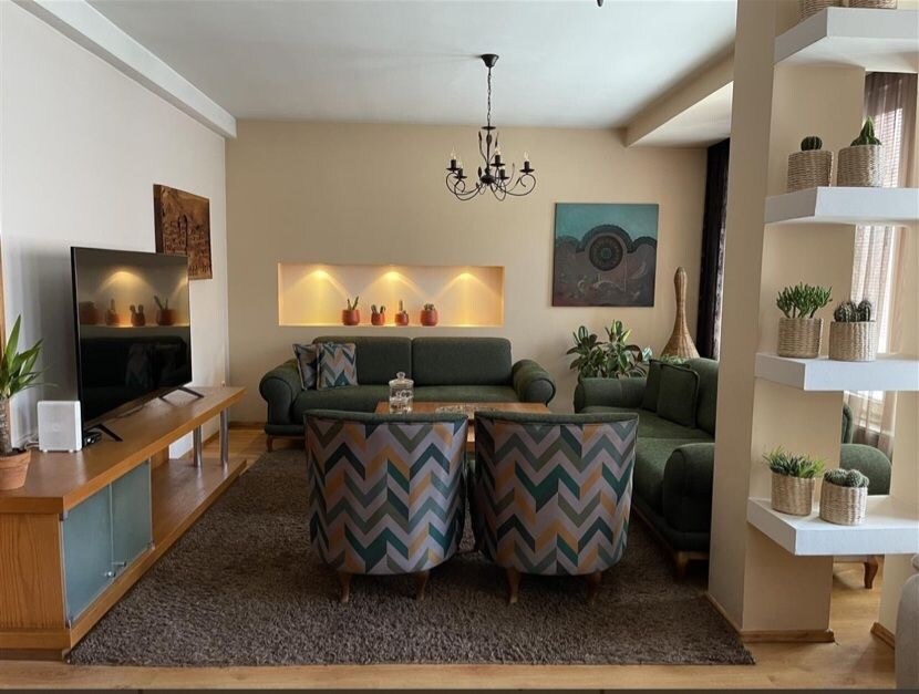 The living area features comfortable green sofas arranged around a stylish coffee table. A large television is mounted on an oak media console. Decorative plants are displayed on shelves, and a textured area rug adds warmth to the space.
