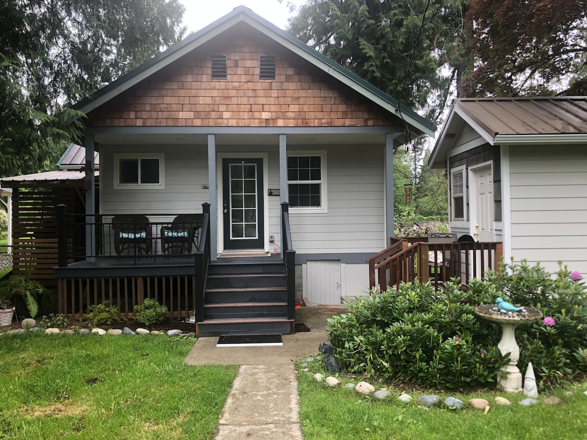 The charming exterior of the house is framed by neatly manicured grass and decorative landscaping. A covered porch features seating arrangements, providing an inviting entrance. The dual-tone siding and shingle roof elements are balanced by natural greenery surrounding the structure.