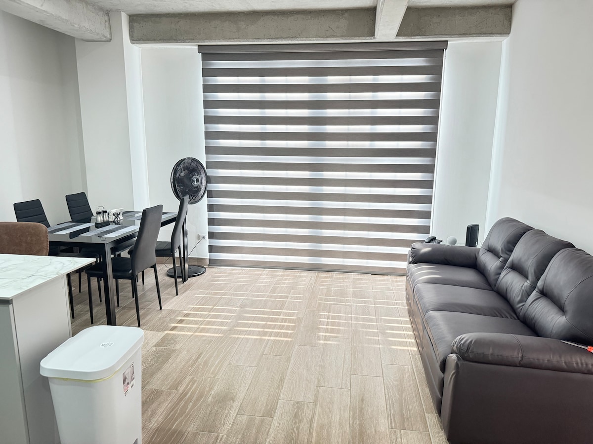 A modern living area features a dark leather sofa positioned alongside a light-toned wall. A sleek dining table with black chairs is visible, complemented by a fan and large window treatment that allows for filtered light to enter the space.