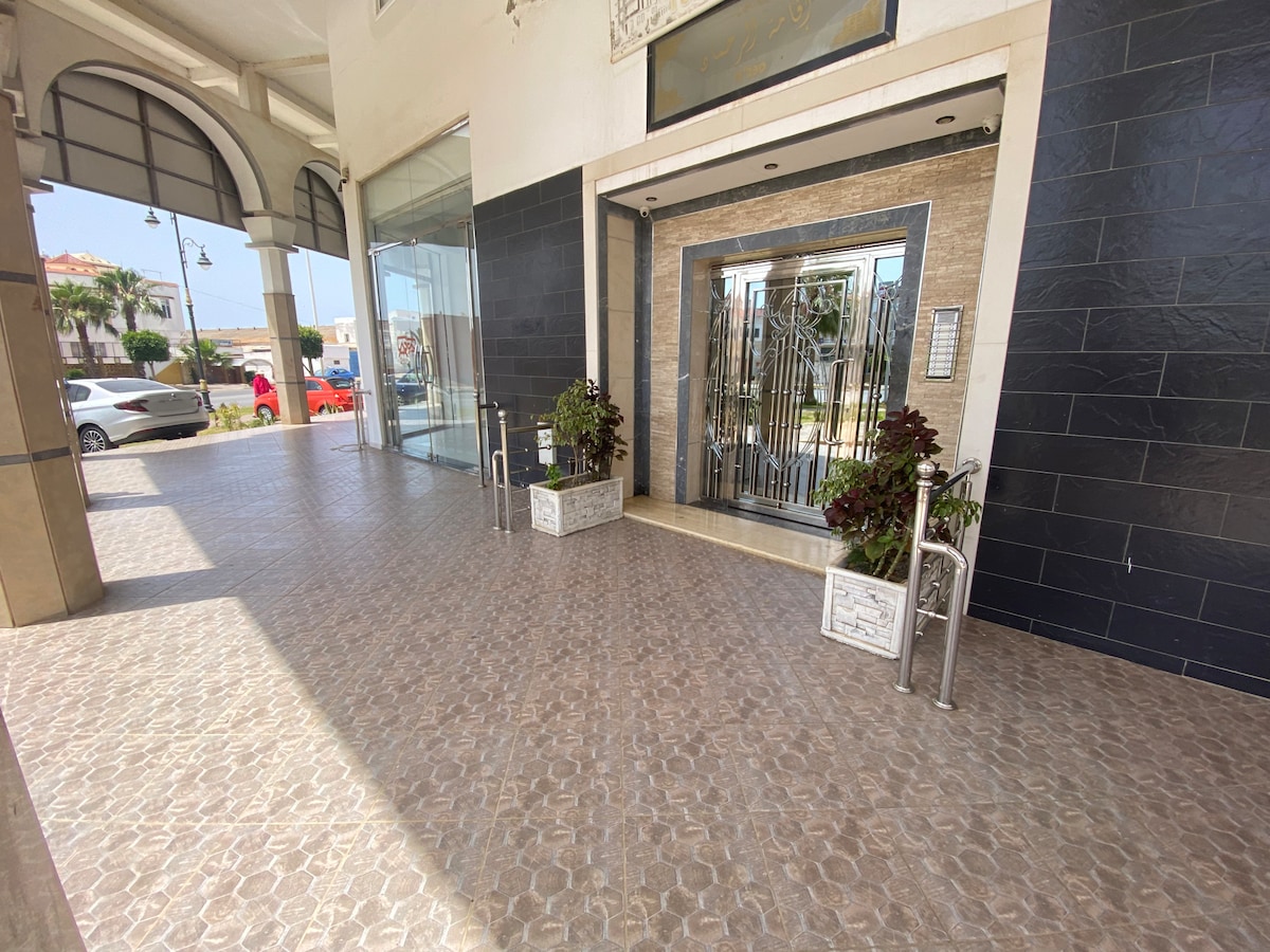 A modern entrance with a sleek design is presented, featuring large glass doors framed by decorative metalwork. Potted plants are placed on both sides of the entrance. The flooring showcases patterned tiles, and natural light brightens the space, while a view of the outside area can be seen.