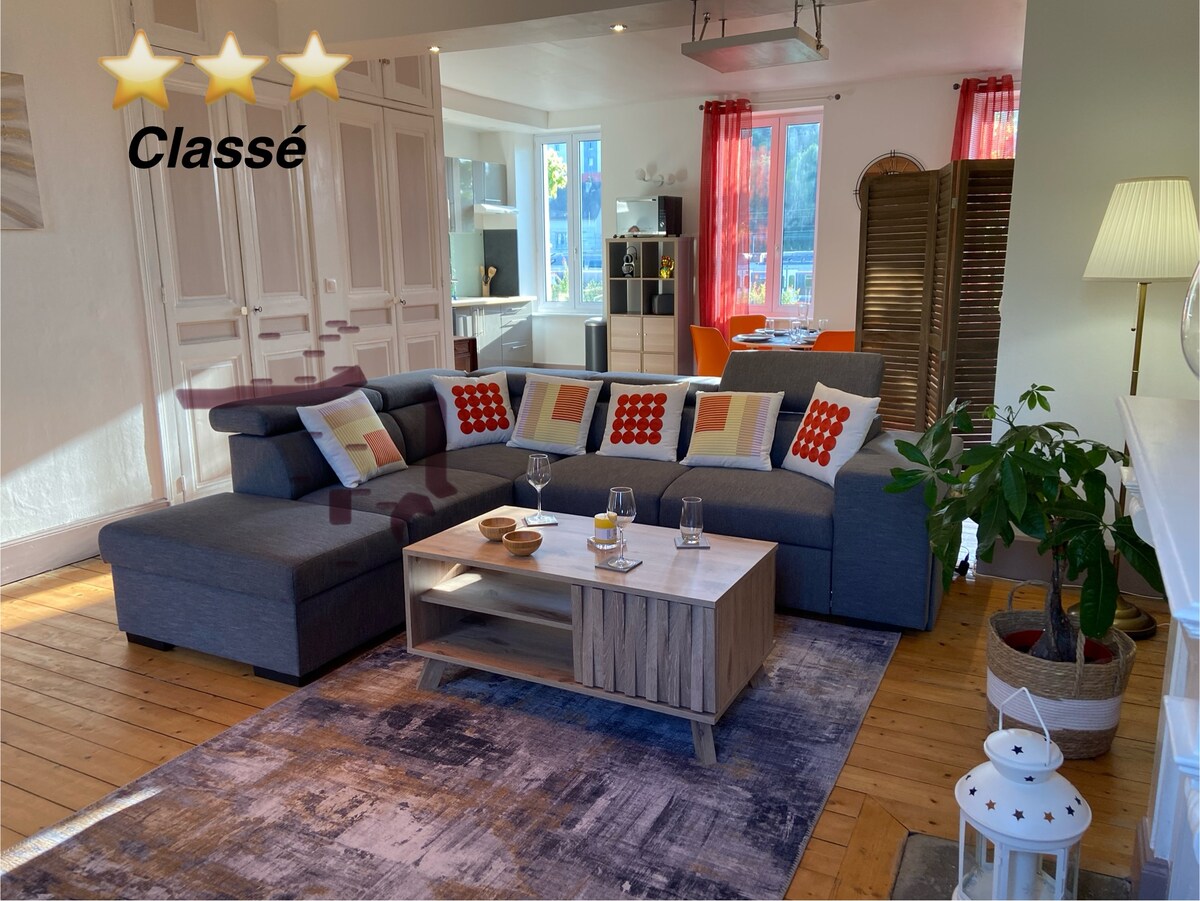 A spacious living area showcases a large grey sectional sofa with colorful cushions and a central coffee table. Natural light floods the room through tall windows, enhancing the warm wooden floors and a subtle area rug design. A dining space is visible in the background.