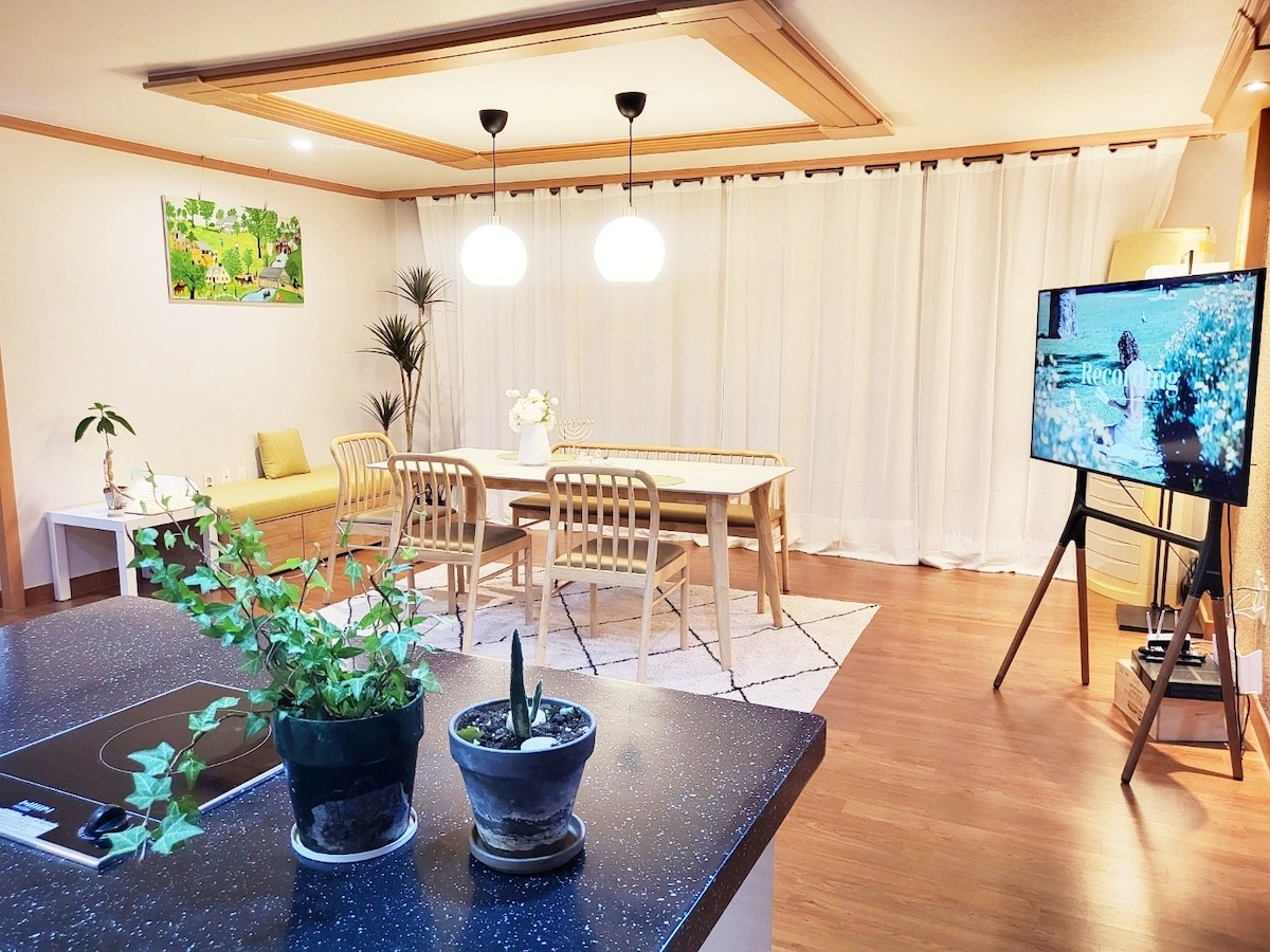 A spacious living and dining area features a large table surrounded by six wooden chairs. Soft natural light filters through sheer curtains, illuminating a cozy seating area with a low sofa. A television stands on an easel, and potted plants add greenery to the space.