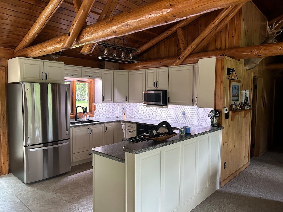 A modern kitchen features sleek cabinetry and a granite countertop. Stainless steel appliances, including a refrigerator and microwave, are positioned for convenience. Wooden beams highlight the ceiling, and a window allows natural light to brighten the space.