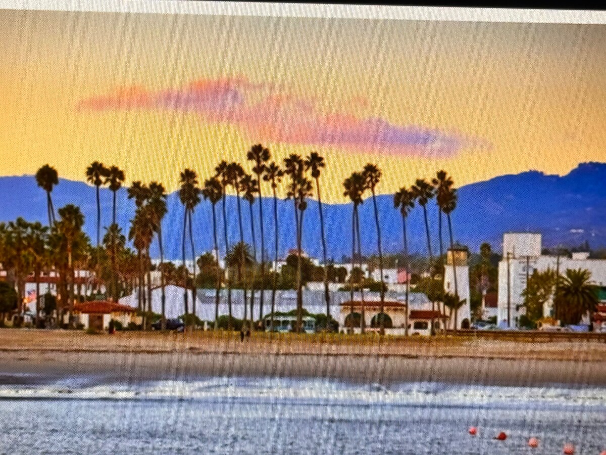 A scenic view captures a coastline lined with palm trees, framed by a backdrop of mountains. The soft colors of the sunset gradient across the sky, reflecting tranquility. The beach is visible in the foreground, with gentle waves lapping at the shore.