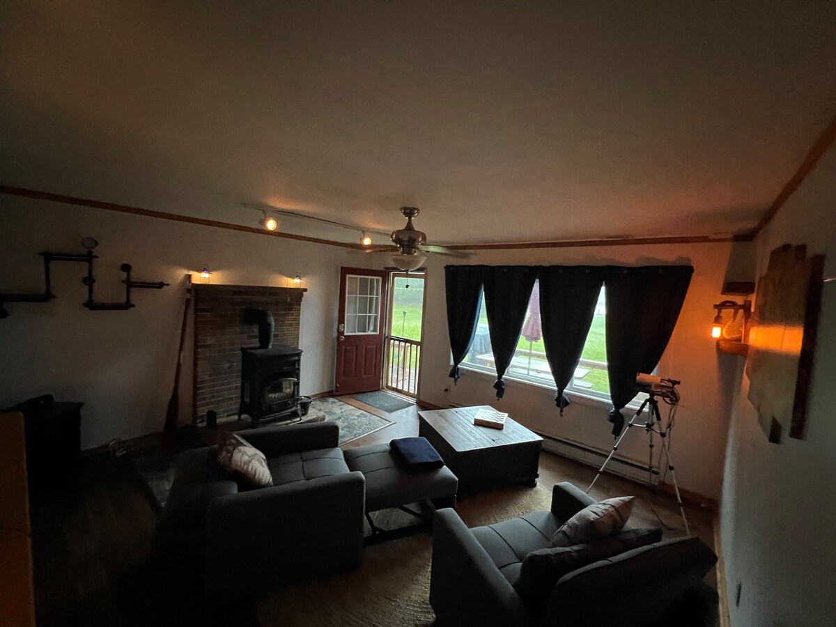 The living room features a wood stove against a brick wall, illuminated by warm lighting. Comfortable seating includes two gray sofas beside a coffee table. Large windows are adorned with dark curtains, bringing in natural light and offering a view of the outdoors.