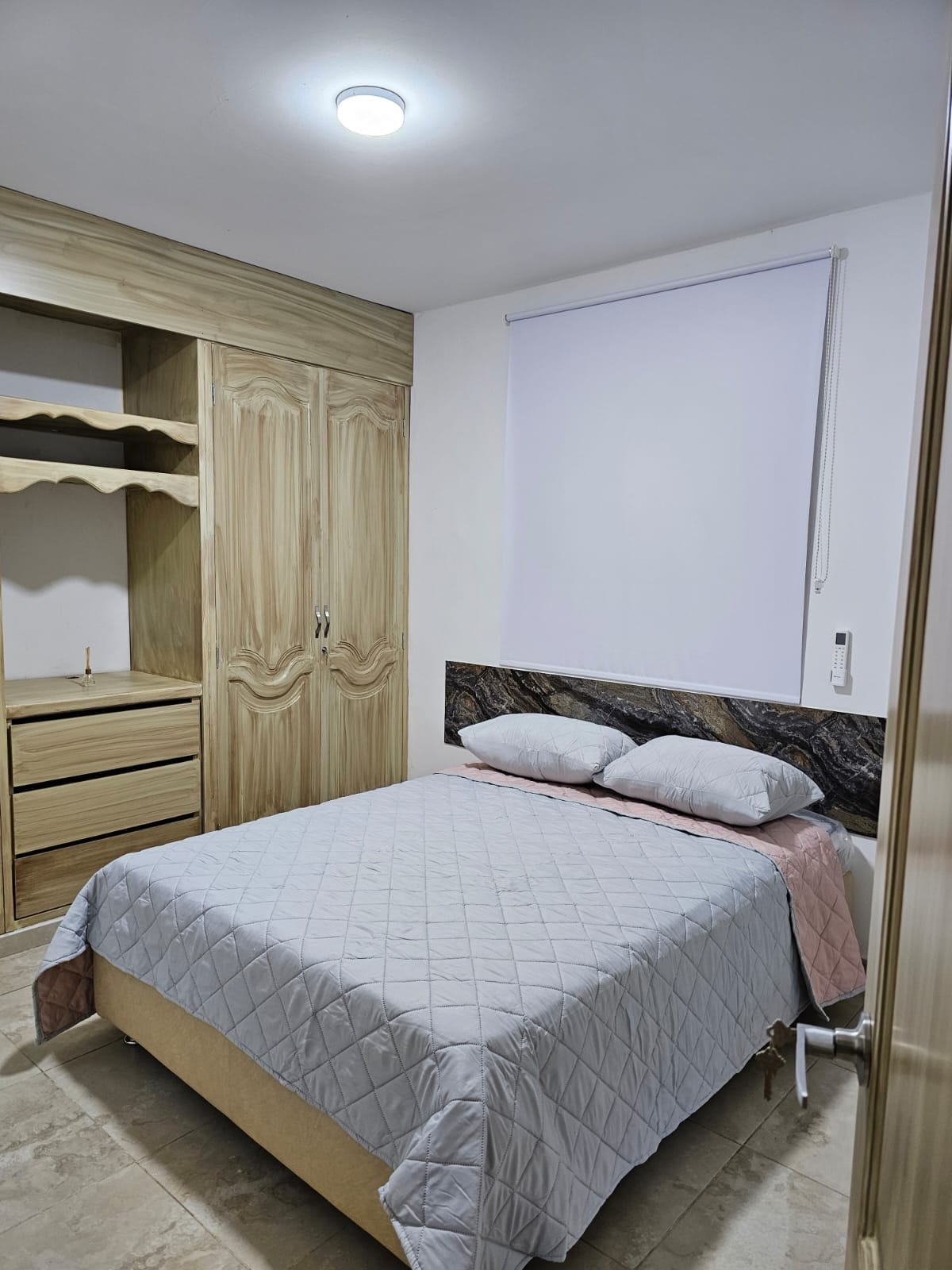A cozy bedroom is featured, equipped with a queen-sized bed covered in a light quilt. Two pillows rest against a decorative headboard. A wardrobe with drawers stands nearby, complemented by a window covered with a light-filtering blind, allowing soft light to enter the space.