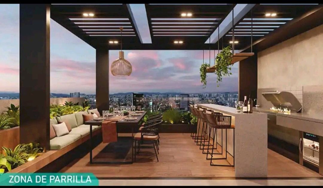 An outdoor dining area is designed with a long table surrounded by chairs, complemented by a built-in barbecue and bar seating. Lush greenery and plants frame the space, while a panoramic city view is captured in the background, illuminated by soft evening light.