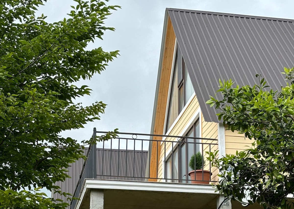 The exterior of a cottage features a modern peaked roof and large windows. A small balcony is visible, adorned with a potted plant, and is framed by lush green trees, enhancing the natural surroundings.