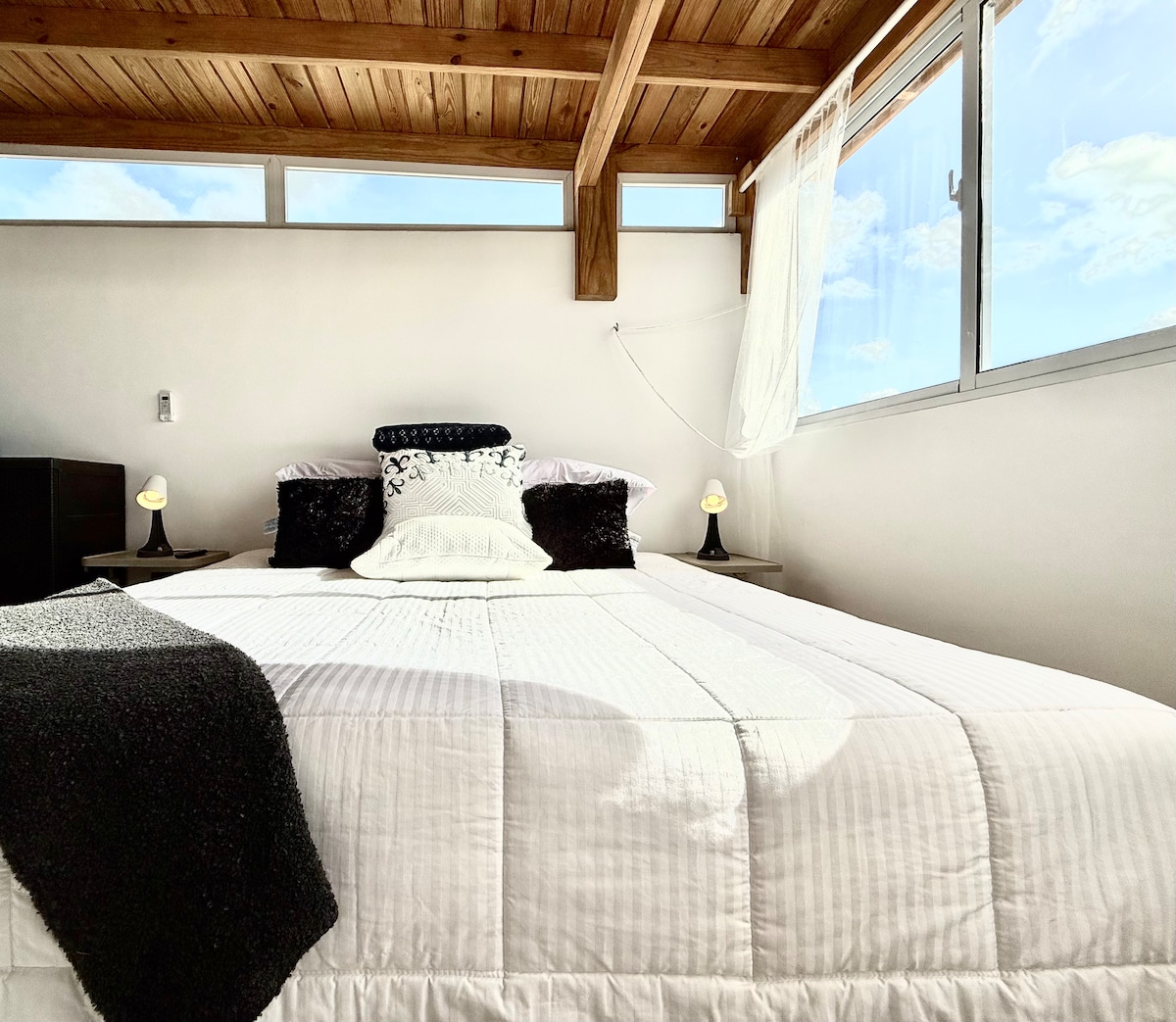 A cozy bedroom features a spacious bed adorned with soft white linens and decorative pillows. The room is illuminated by natural light streaming through large windows, with wooden beams enhancing the warm ambiance. Two bedside lamps are positioned on either side of the bed.