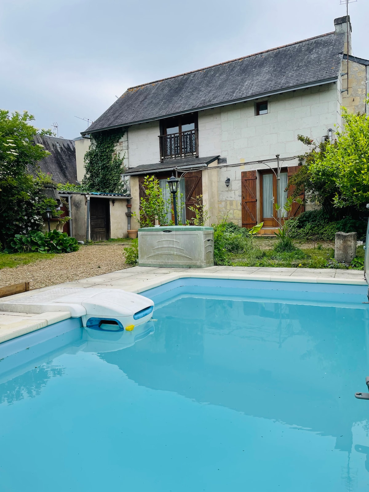 A serene swimming pool reflects the surrounding greenery, bordered by smooth stone edges. The house features multiple wooden shutters, partially open, and a gravel pathway leads to the entrance. Lush plants and trees create a natural ambiance around the outdoor space.