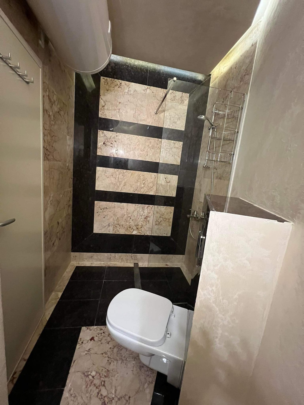 The bathroom features a modern design with marble-patterned walls and sleek black flooring. A toilet is positioned against one side, while a glass shower is visible with a rainfall showerhead. Chrome fixtures add a polished touch to the overall aesthetic.