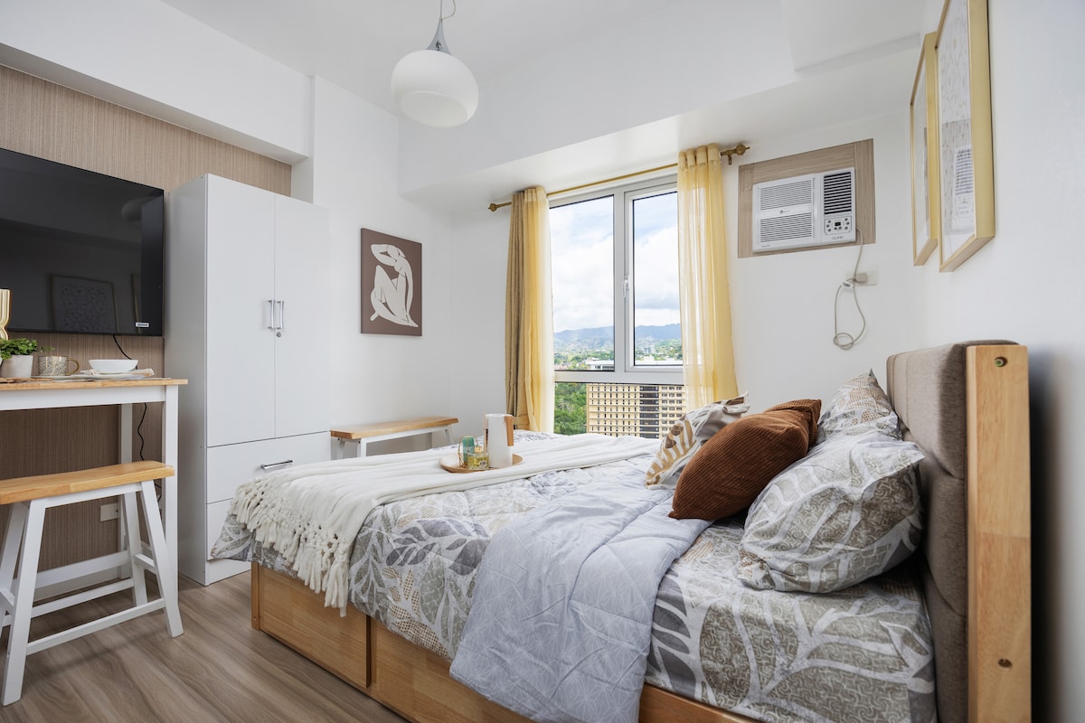 The studio unit features a spacious bed dressed in neutral tones, complemented by decorative pillows. A wooden dining table is visible in the background, alongside a television mounted on the wall. Natural light fills the room through a large window with sheer curtains, enhancing the cozy atmosphere.