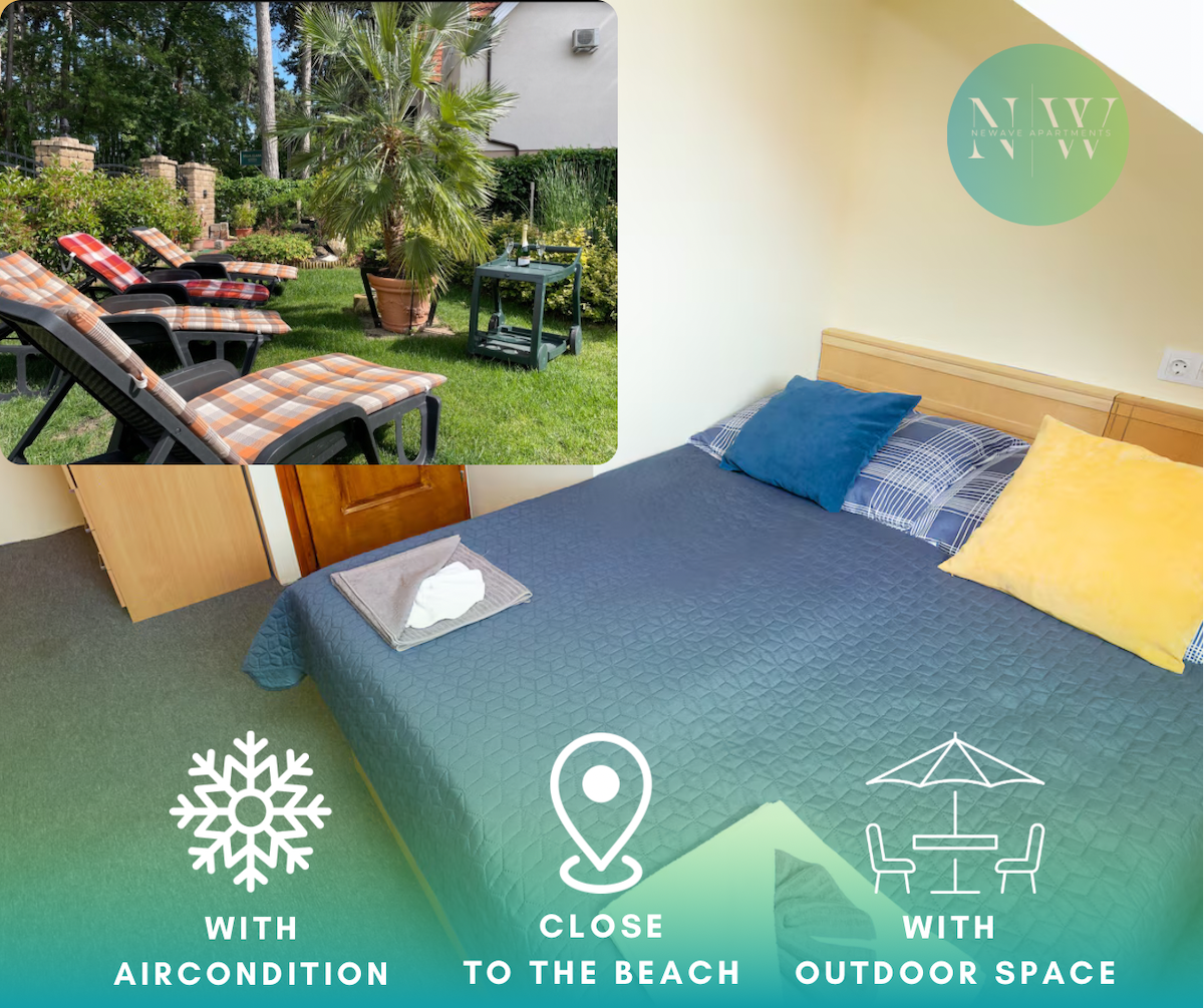 A cozy sleeping area features a spacious bed topped with a blue and white blanket, accompanied by several colorful pillows. Sunlight enters through a nearby window, illuminating a lush outdoor space visible through the window, which includes lounge chairs and greenery.