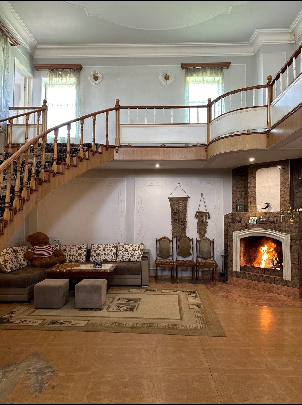 A spacious living area features a curved staircase leading to an upper level. Soft seating arrangements include a sectional couch with patterned cushions, complemented by a stone fireplace that provides warmth. Decorative wall hangings and three chairs enhance the inviting ambiance.