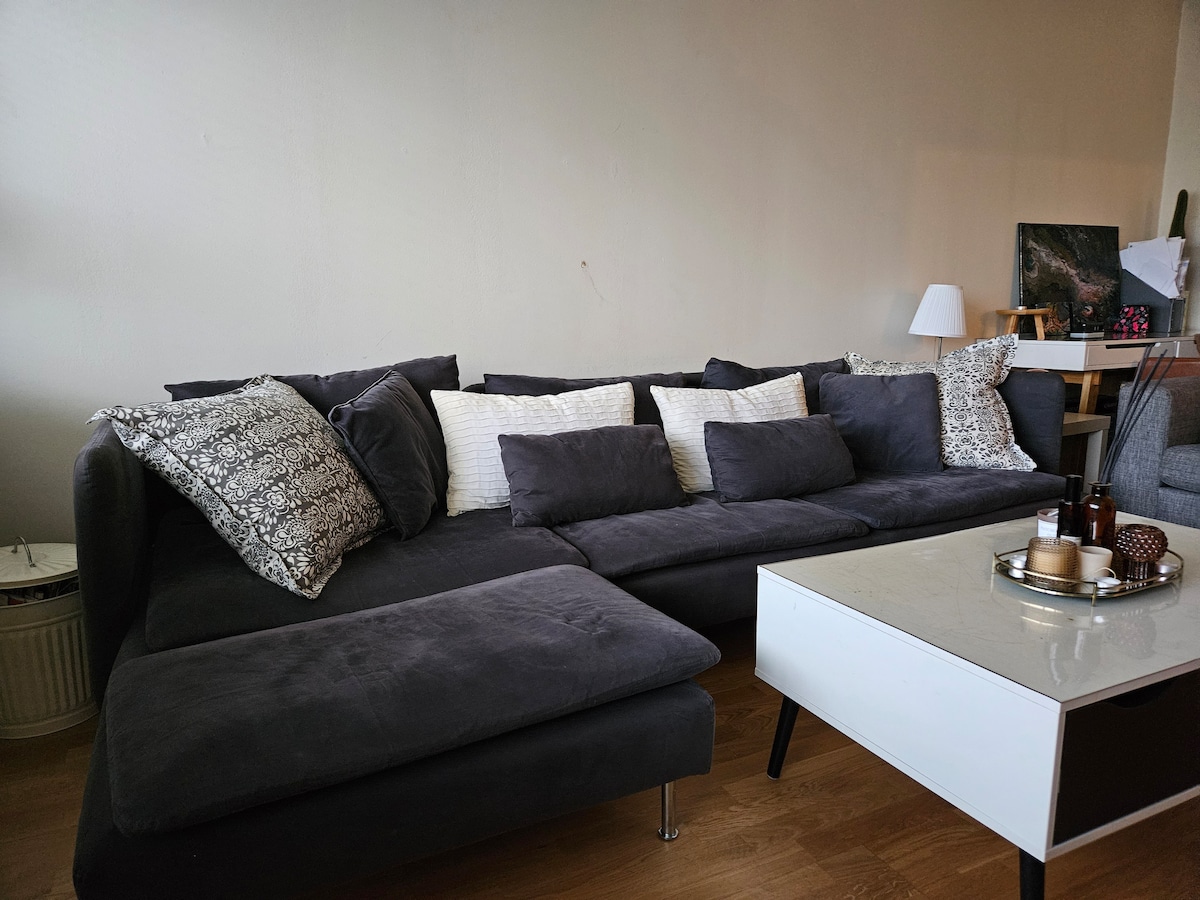 A spacious, dark grey sectional sofa is featured, adorned with decorative pillows in contrasting tones. A coffee table with a light-colored surface sits in front, displaying candles and a small decorative tray. A lamp is positioned nearby, providing subtle lighting.
