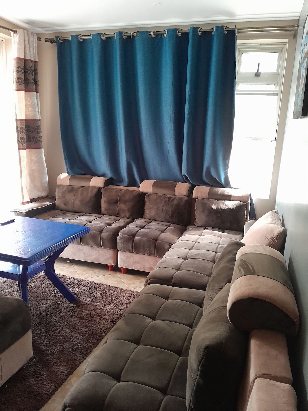 A spacious living area features a large sectional sofa with contrasting brown and beige upholstery, arranged around a blue coffee table. Light filters through large windows, complemented by long, textured curtains in shades of blue and beige, adding depth to the room.