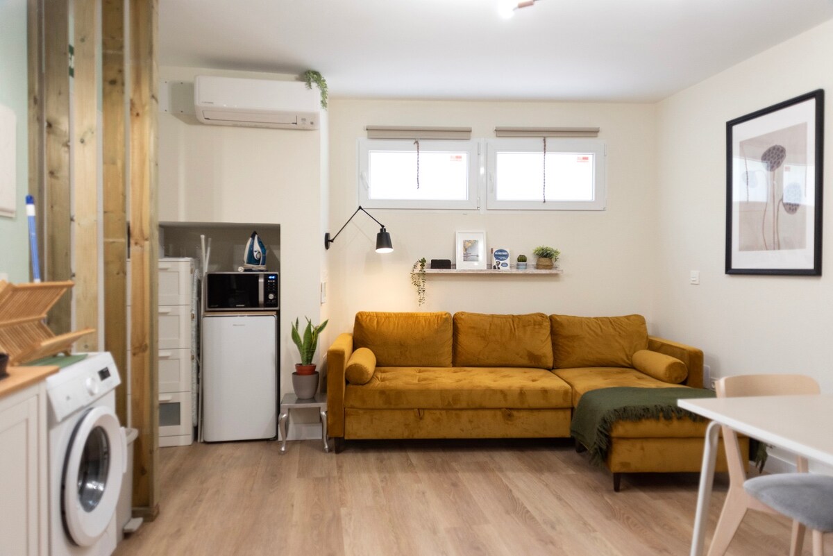 A cozy living space features a yellow sofa complemented by a green throw. Above, a wooden shelf is lined with decorative items. A compact kitchenette is visible, containing essential appliances, while a washing machine is integrated into the space. Natural light enters through two windows.