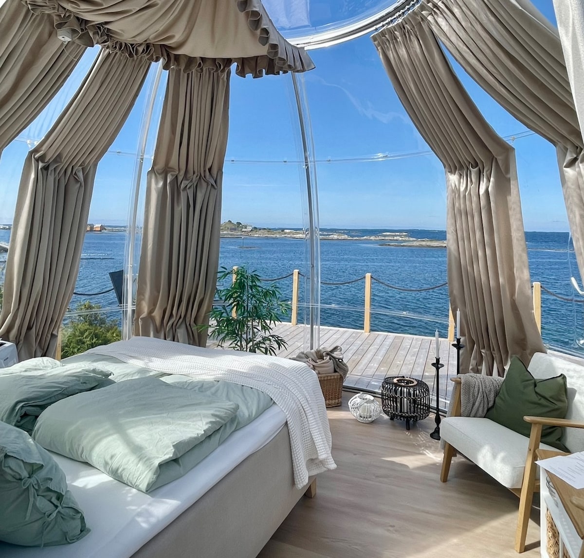 A clear dome interior reveals a cozy sleeping area with light green bedding atop a spacious bed. Soft curtains frame large openings, allowing natural light to illuminate the room. A plant adds a touch of greenery, and the expansive view of the sea is visible beyond a private deck.