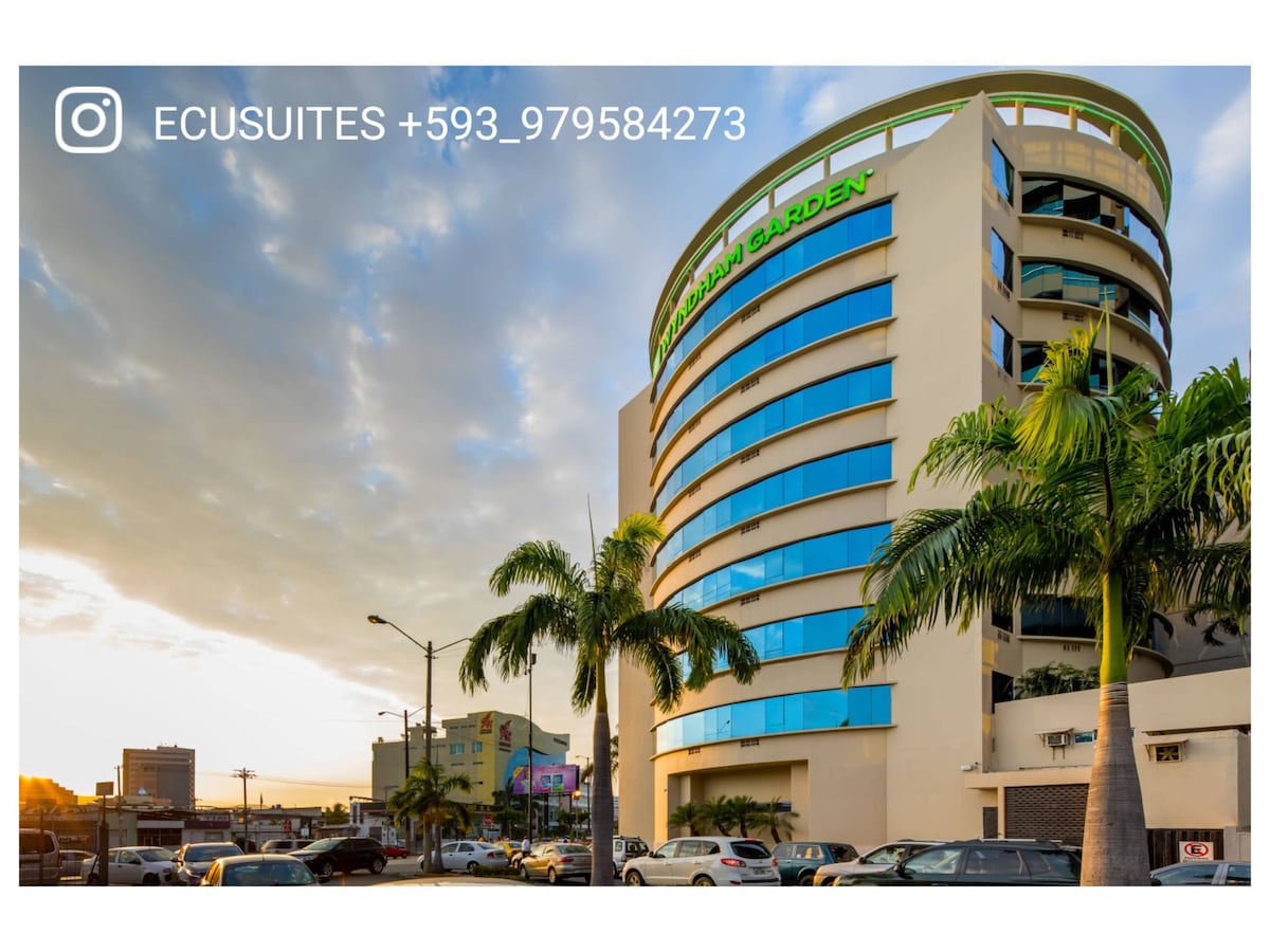 The exterior of a modern building is displayed, featuring a circular design with large windows. The façade is highlighted by greenery from palm trees and the hotel's name is visible in bright green lettering. Parking areas and surrounding streets offer a bustling atmosphere.