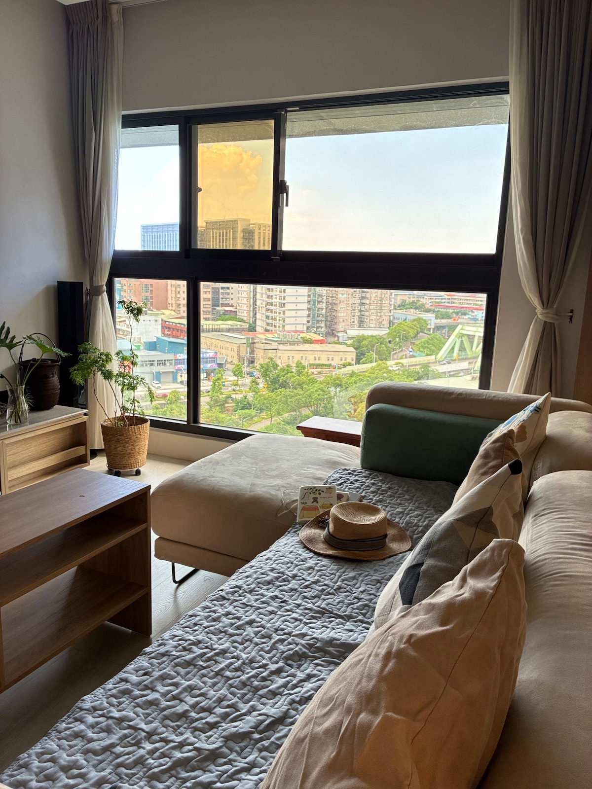 A comfortable seating area is shown featuring a light-colored sofa with decorative pillows. A large window offers a view of the surrounding residential area and cityscape, while greenery is visible outside, bringing a sense of openness and natural light into the space.