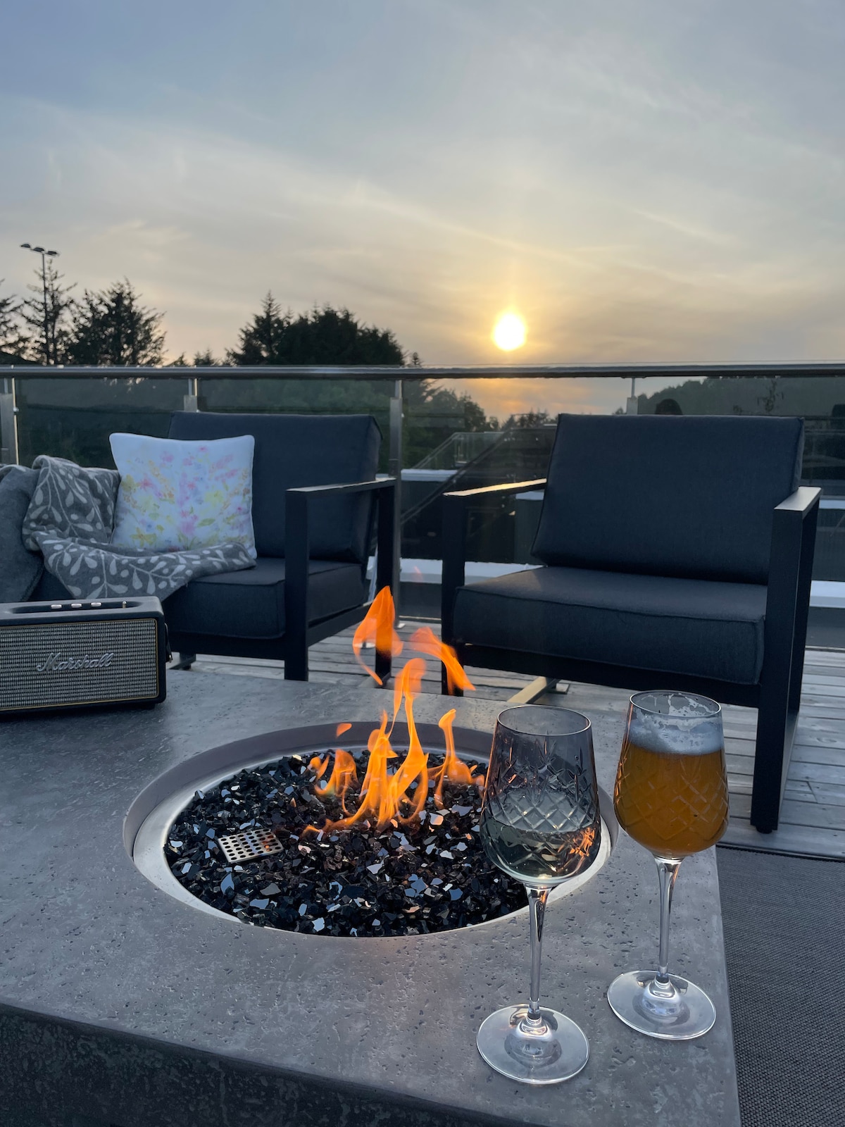 An outdoor seating area features two modern chairs positioned beside a fire pit, with flames flickering above a bed of decorative stones. Two glasses, one filled with sparkling water and the other with a light beverage, sit on the table, capturing an evening ambiance with a sunset in the background.