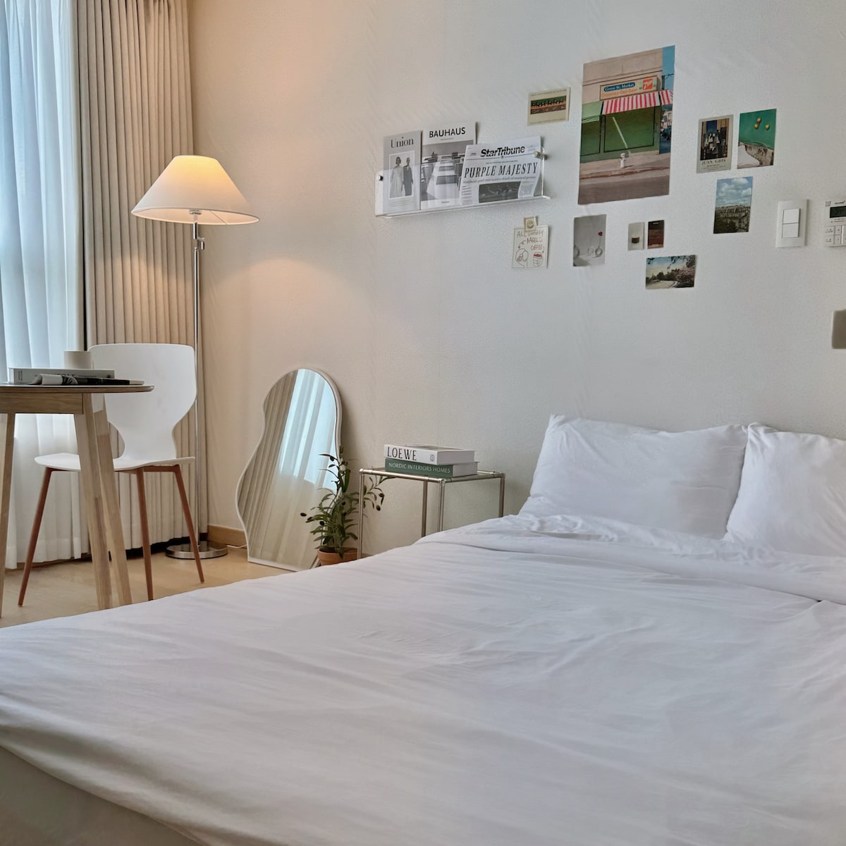 A bright and airy room features a spacious bed with crisp white linens. A minimalist table and a chair sit nearby, complemented by a decorative mirror. Walls are adorned with various photographs, with natural light streaming in through sheer curtains.