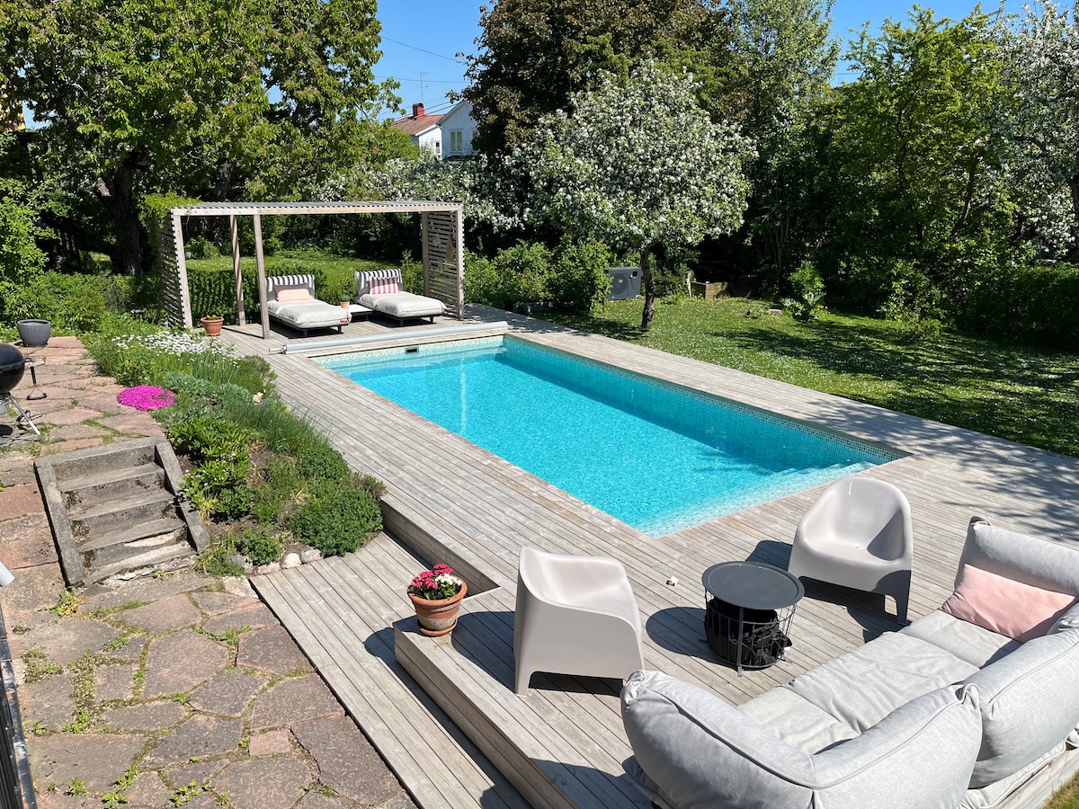 A serene outdoor space features a swimming pool surrounded by wooden decking. Two lounge chairs are positioned nearby, accompanied by a cozy seating area with plush sofas. Potted plants add a touch of greenery, and the lush garden provides a tranquil backdrop.
