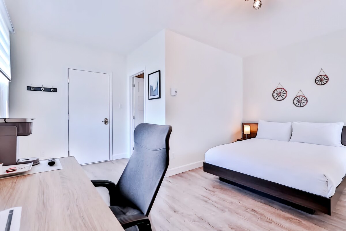 A spacious room is presented with a large bed and a modern desk. A comfortable chair faces a window, allowing natural light to brighten the area. Decorative wall hangings add subtle character, while sleek furnishings provide a clean and organized atmosphere.