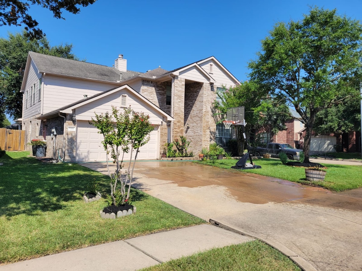 The exterior of a spacious two-story home is showcased, featuring a combination of stone and light-colored siding. A well-maintained lawn surrounds the property, complemented by trees and shrubs. A driveway leads to a two-car garage, with a basketball hoop visible on one side.
