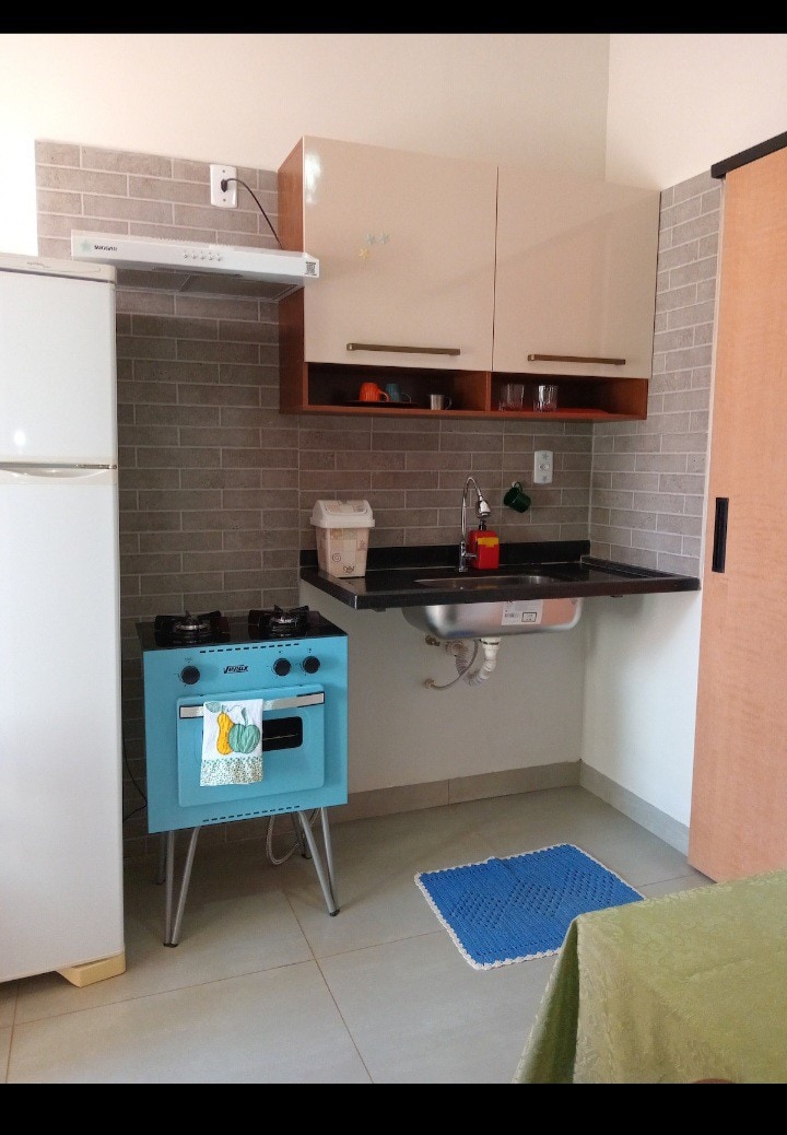 A compact kitchen area is equipped with essential appliances, including a refrigerator and a blue stove with four burners. Upper cabinets provide storage, and a sink is positioned beneath a wall-mounted shelf. A textured mat lies on the floor near the sink, complementing the modern design.