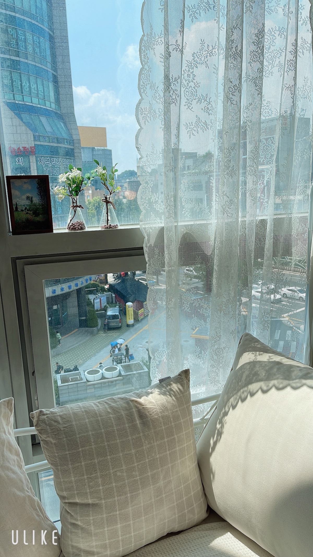 A close-up of a window reveals delicate lace curtains gently filtering sunlight. Two small decorative vases are placed on the windowsill. Outside, a cityscape is visible, with people and vehicles creating a lively atmosphere.