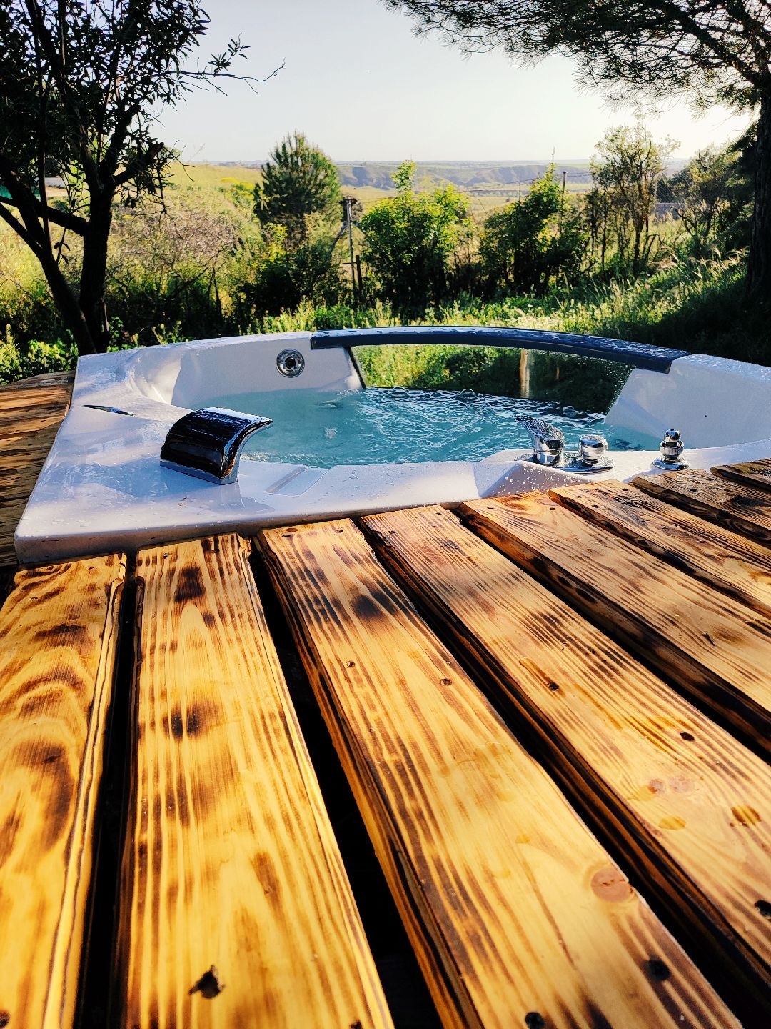 A private jacuzzi is nestled on a wooden deck, surrounded by lush greenery and offering mountainous views. The clear water glistens under the sunlight, inviting relaxation in a serene natural setting.
