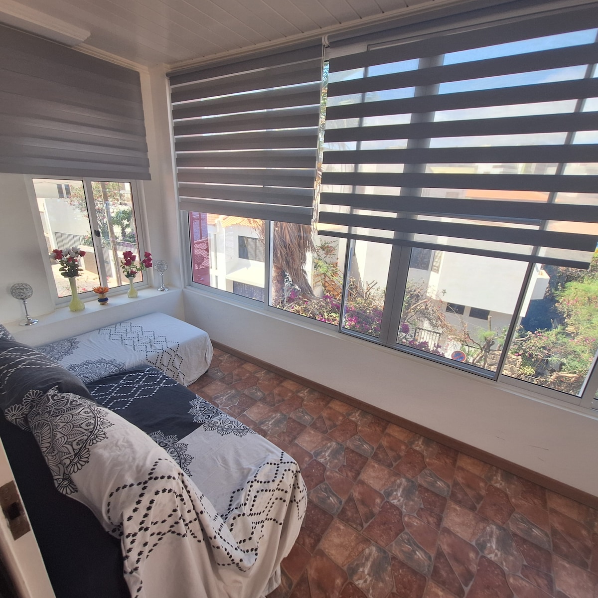 A well-lit sleeping area features two beds positioned near large windows adorned with sleek, striped shades. Light flows in, revealing a vibrant view of the outside greenery, while decorative pillows and bedding add subtle texture to the space.